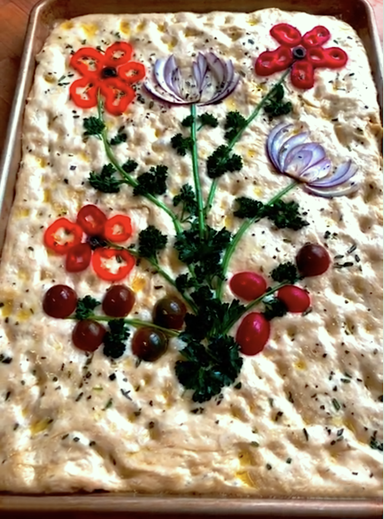 A fresh baked focaccia topped with vegetables arranged to look like a flower bouquet.