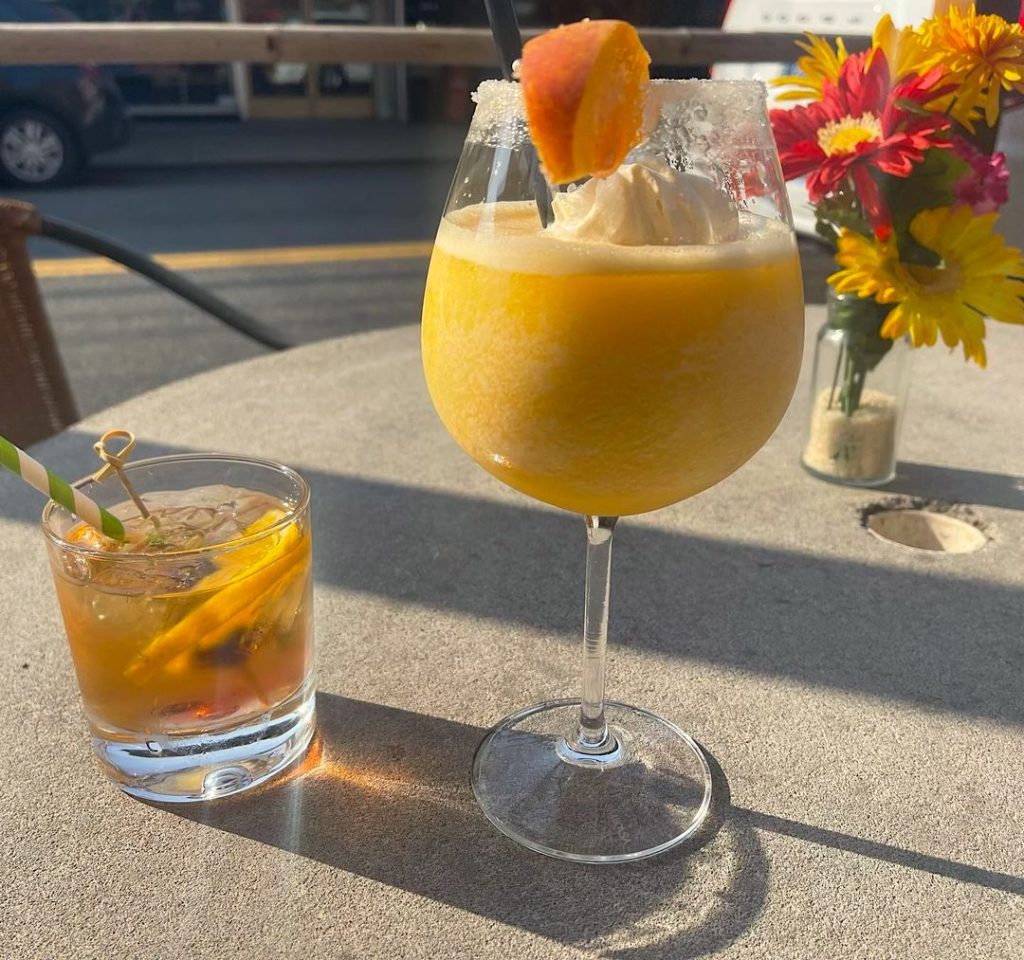 A sunny yellow peach margarita on a patio table with some pretty daisies and a whiskey on the side.